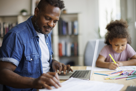 father working on laptop while daughter colors in the background